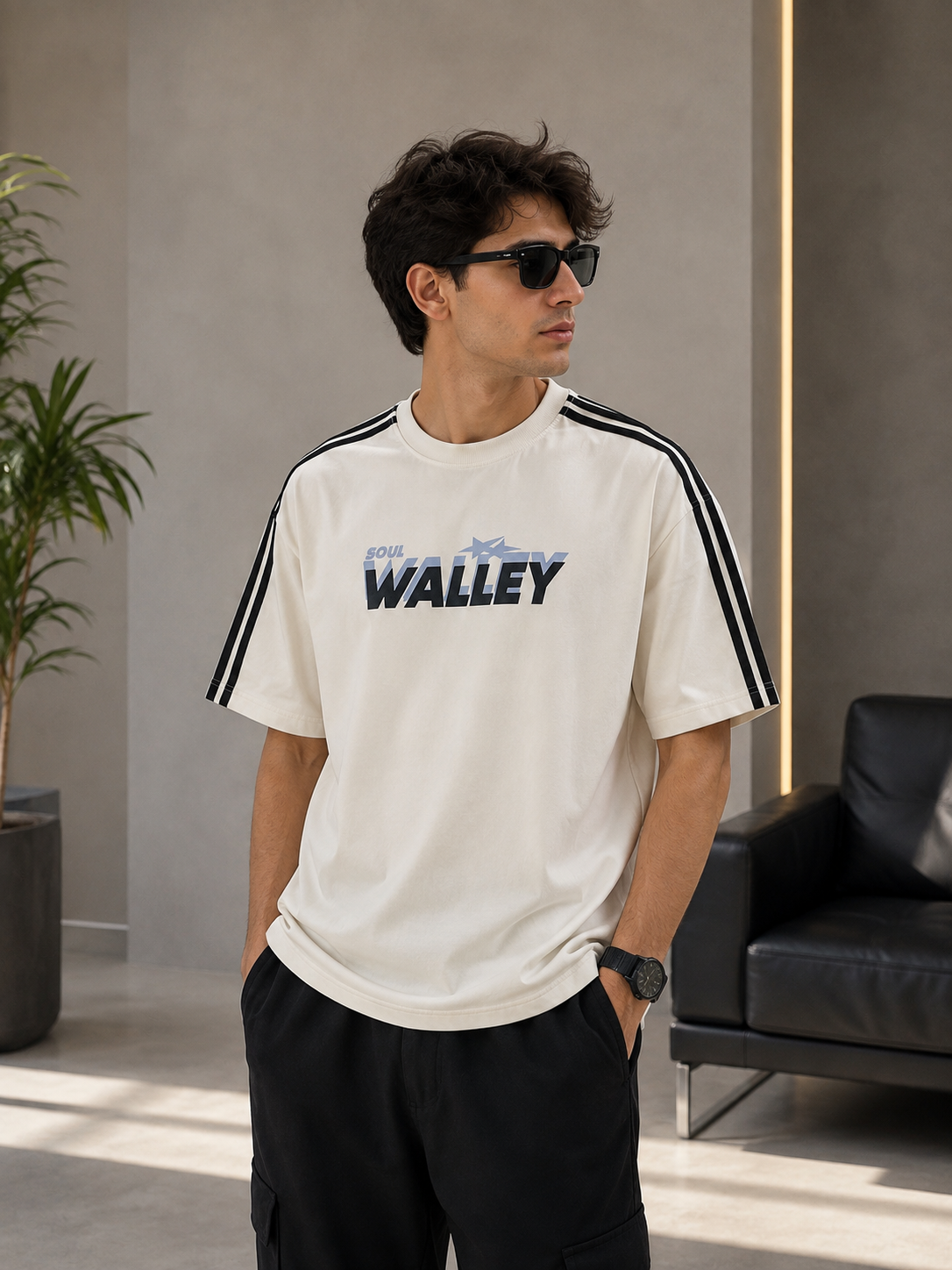 A man wearing a cream-colored short-sleeve t-shirt with black stripes on the shoulders and a blue 'WALLEY' logo on the chest, paired with black pants, standing in a modern indoor setting.