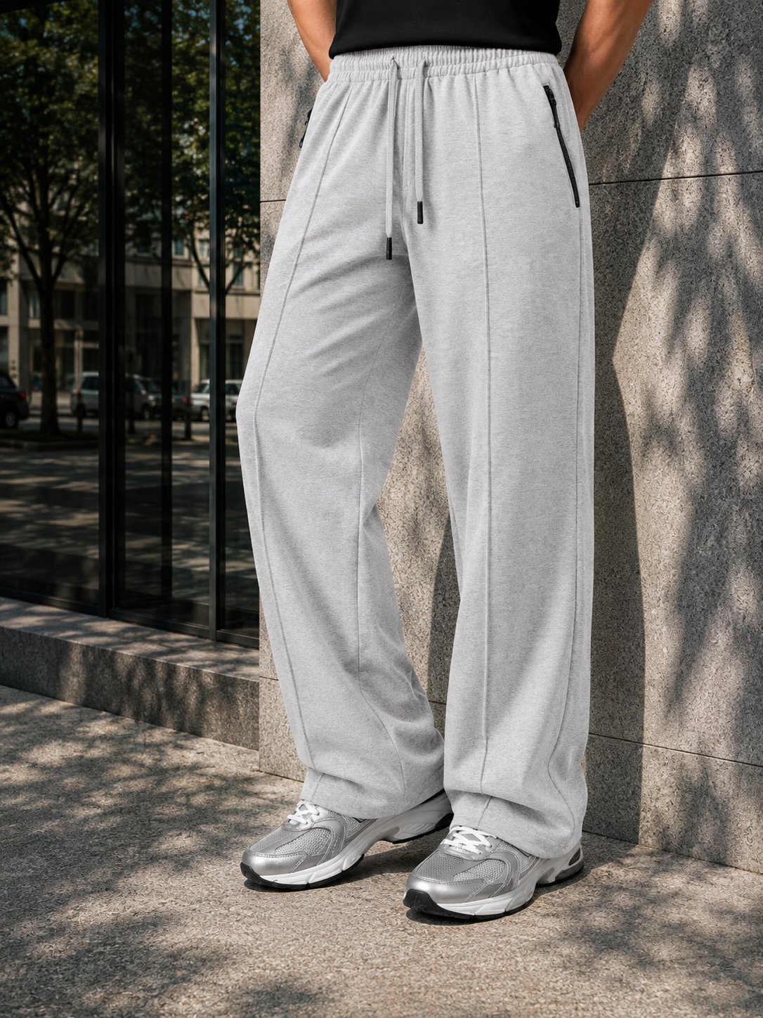 A person wearing light gray wide-leg sweatpants with a drawstring waist and zippered side pockets, paired with silver sneakers, standing outdoors against a stone wall.