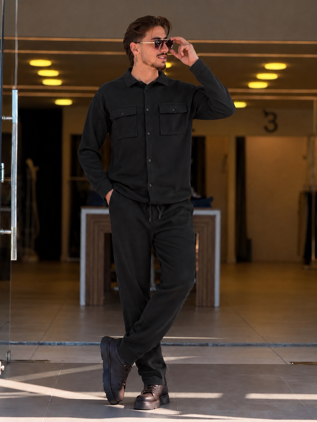 A man wearing a black long-sleeve button-up shirt and matching black pants, styled with dark brown leather shoes and sunglasses, standing in a modern indoor hallway.