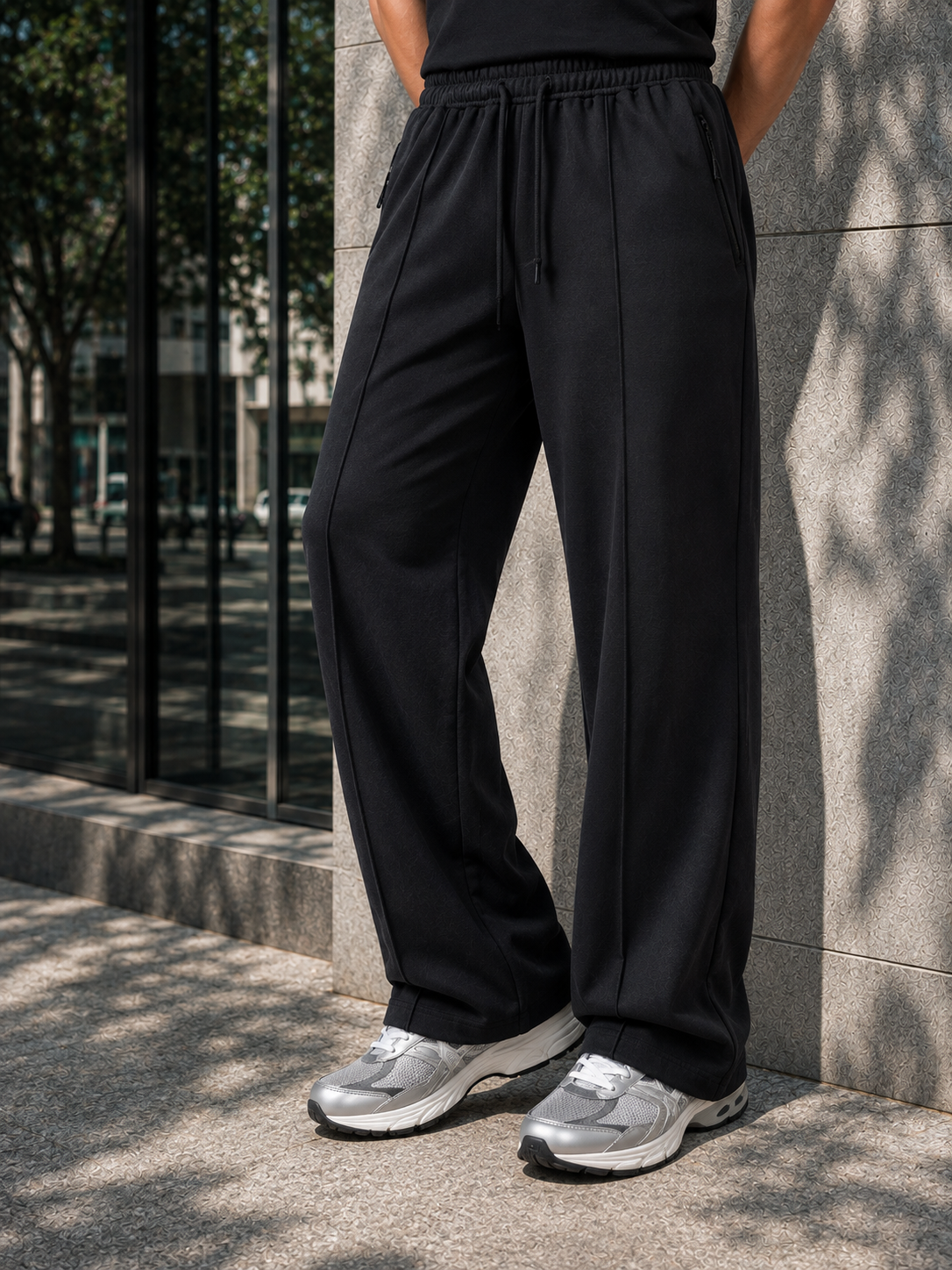 A person wearing black wide-leg drawstring sweatpants and silver sneakers, standing outdoors against a gray stone wall with tree shadows.