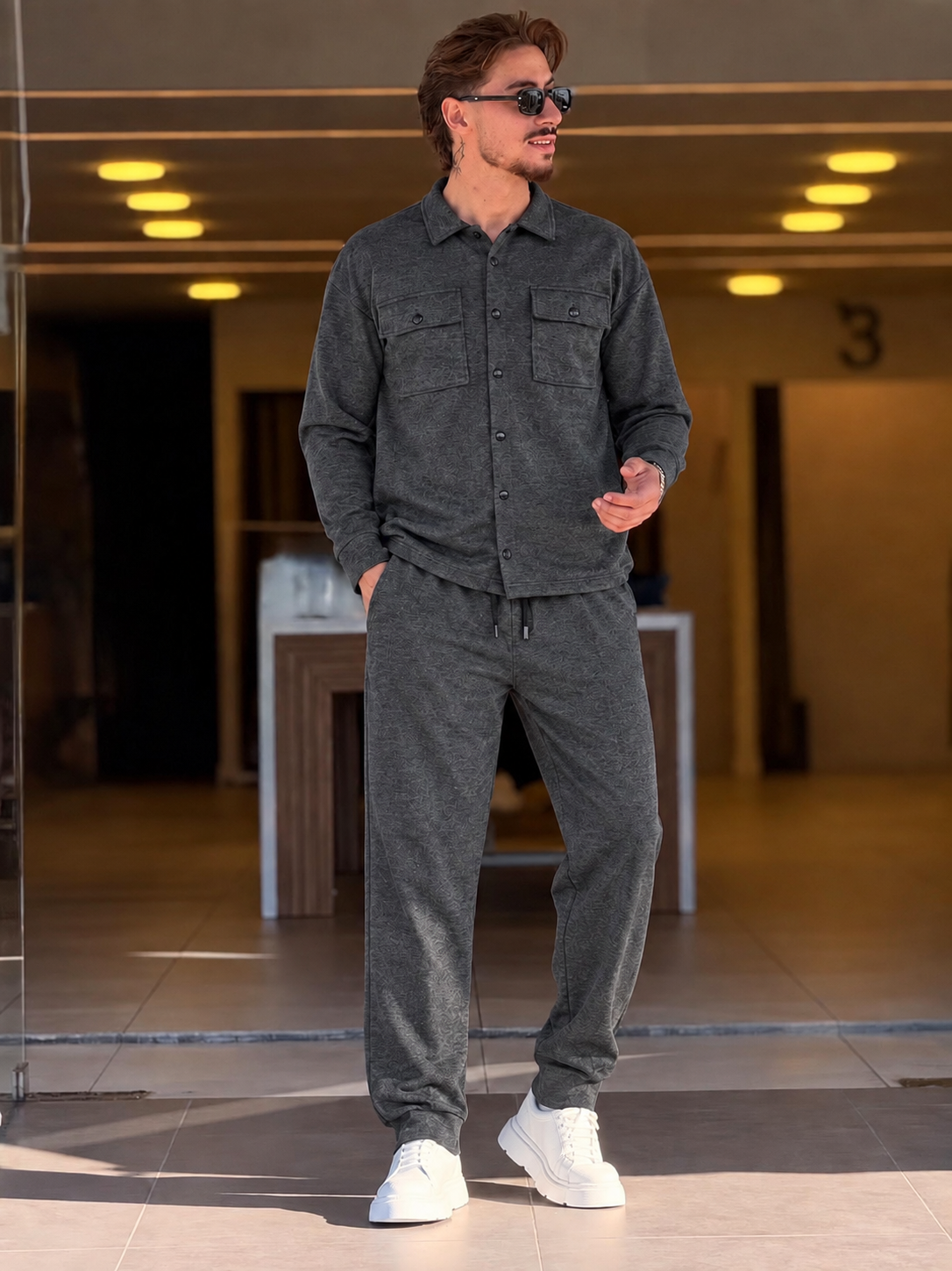 A man wearing a dark gray two-piece lounge set consisting of a long-sleeve button-up shirt and matching jogger pants, paired with white sneakers, standing in a modern indoor lobby.