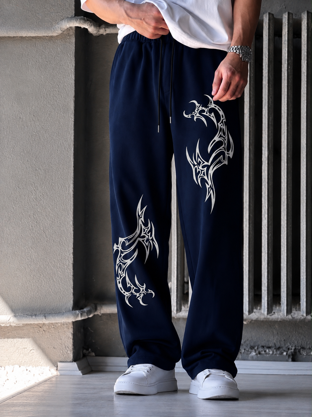 A person wearing navy blue sweatpants with large white tribal-style graphic prints on both legs, paired with a white t-shirt and white sneakers, standing against a concrete wall and radiator.