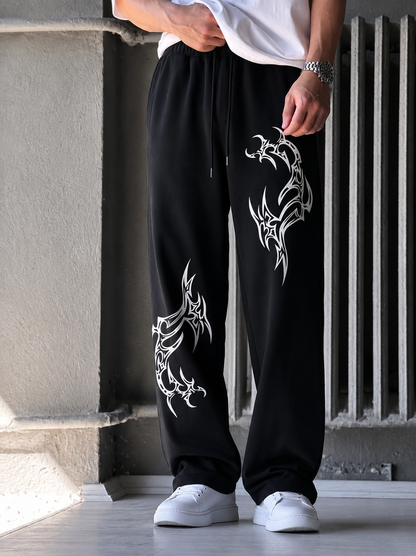 A person wearing black drawstring sweatpants with large white tribal-style graphic prints on both legs, paired with a white t-shirt and white sneakers, standing in front of a concrete wall and radiator.