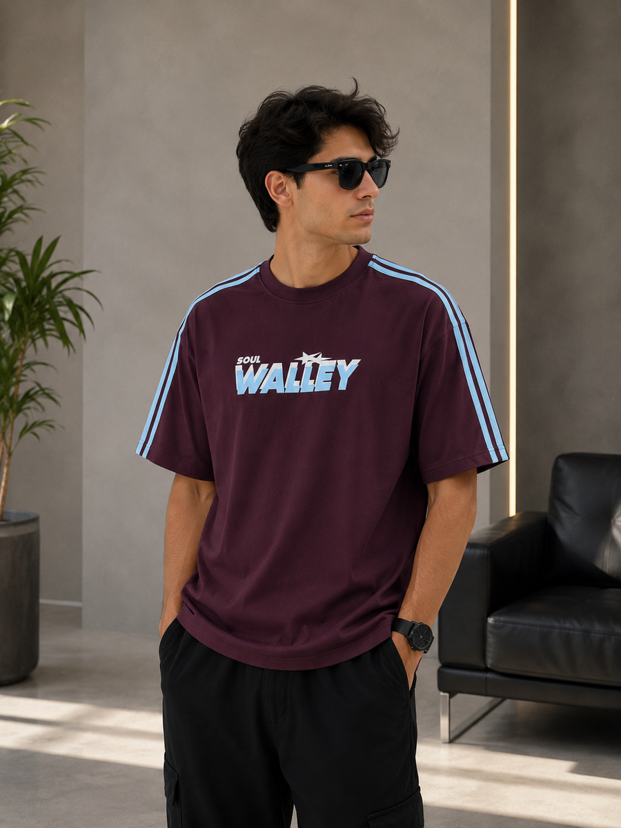 A man wearing a maroon short-sleeve t-shirt with 'THE WALLEY' printed in light blue across the chest, styled with black cargo pants and sunglasses in a modern indoor setting.
