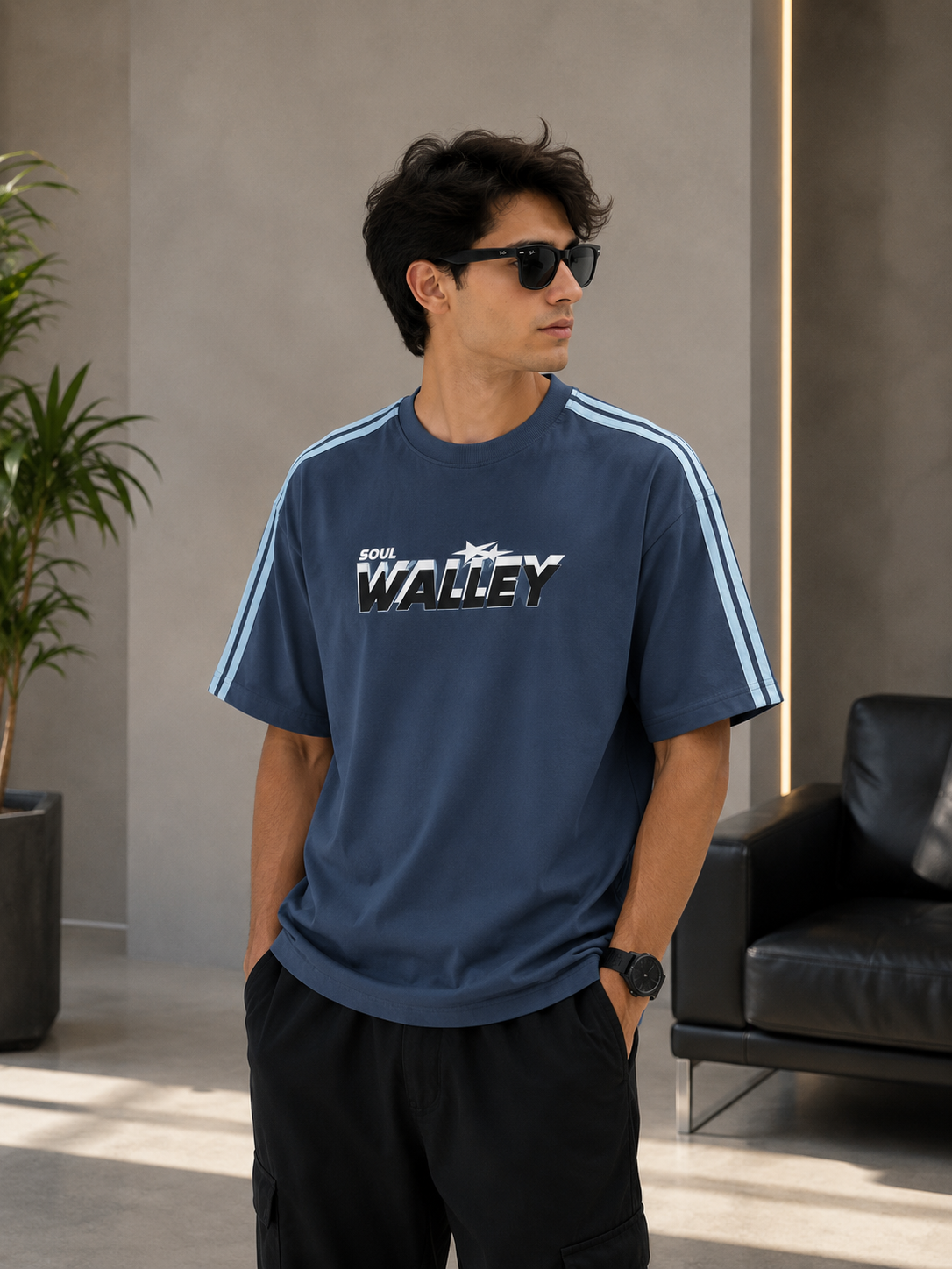 A man wearing a navy blue short-sleeve t-shirt with 'WALLEY' printed across the chest and white stripes on the shoulders, paired with black pants, standing in a modern indoor setting.