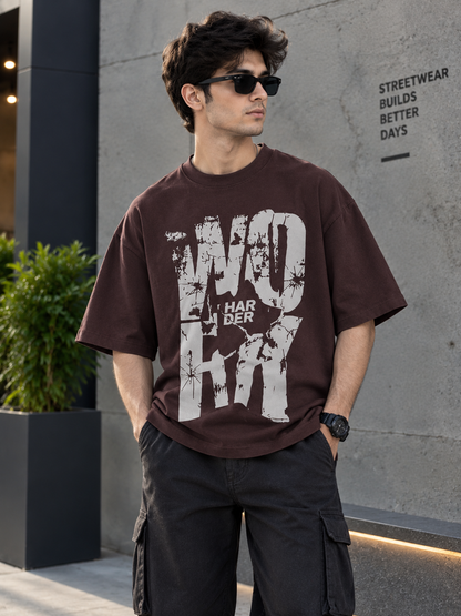 A man wearing a dark brown oversized graphic t-shirt with a distressed white print that reads 'WORK HARDER', paired with black cargo pants and sunglasses, standing in front of a modern concrete wall.