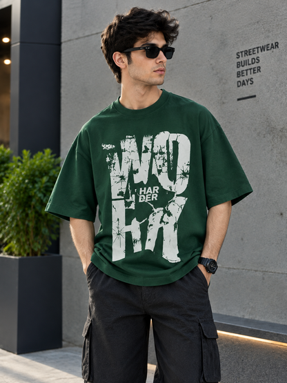 A man wearing a dark green oversized t-shirt with a distressed white graphic print that reads 'WORK HARDER', paired with black cargo pants and sunglasses, standing outdoors against a gray concrete wall.