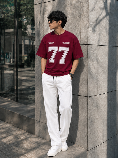 A man wearing a burgundy t-shirt with a large white '77' graphic and white sweatpants, leaning against a gray stone pillar outdoors.