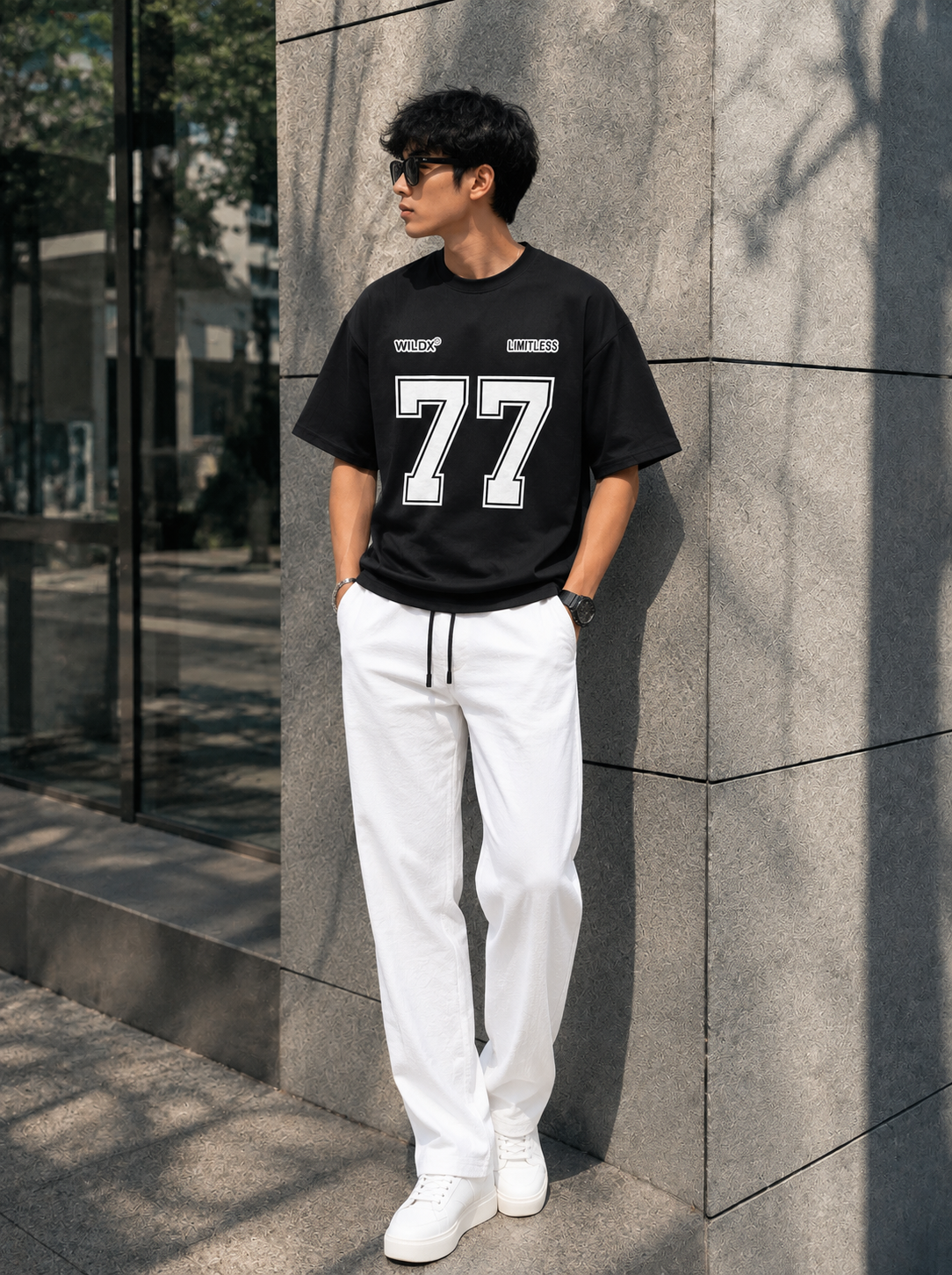 A man wearing a black short-sleeve t-shirt with a bold white '77' graphic on the front, paired with white drawstring sweatpants and white sneakers, leaning against a gray stone wall outdoors.