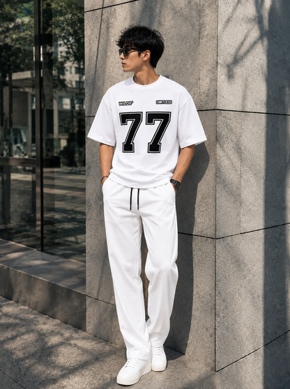 A man wearing a white crewneck t-shirt with a large black '77' graphic and white sweatpants, standing outdoors against a gray stone wall.