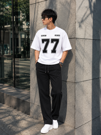 A man wearing a white short-sleeve t-shirt with a large black '77' graphic on the front, paired with black drawstring sweatpants and white sneakers, standing outdoors against a gray stone wall.