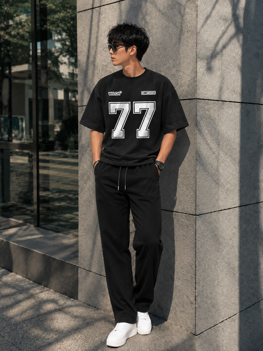 A man wearing a black short-sleeve t-shirt with a large white '77' graphic on the front, black drawstring sweatpants, and white sneakers, standing against a gray stone wall.