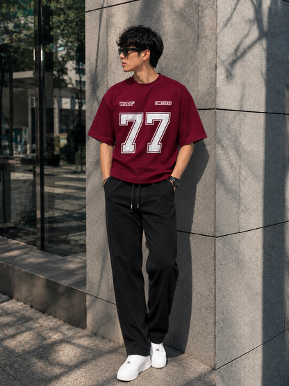 A man wearing a burgundy t-shirt with a large white '77' graphic and black sweatpants, standing outdoors against a gray stone wall.