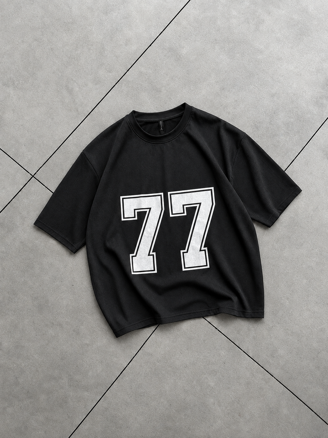 A black short-sleeve t-shirt with a large white '77' graphic printed on the front, laid flat on a gray tiled floor.