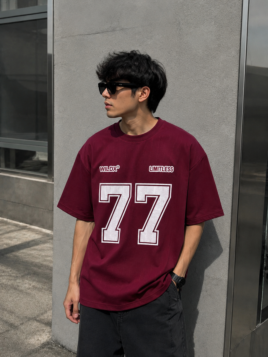 A man wearing an oversized maroon t-shirt with 'WILDFOX' and 'LIMITLESS' printed on the chest, alongside a large white '77' graphic, standing against a gray concrete wall.