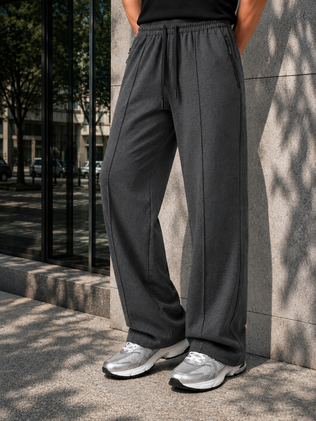 A person wearing dark gray wide-leg drawstring sweatpants and silver sneakers, standing outdoors against a stone wall with tree shadows.