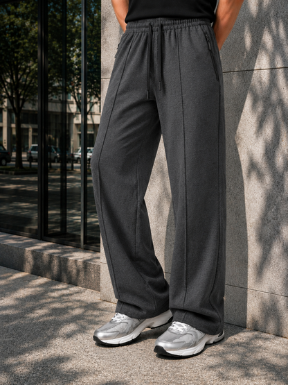 A person wearing dark gray wide-leg drawstring sweatpants and silver sneakers, standing outdoors against a stone wall with tree shadows.