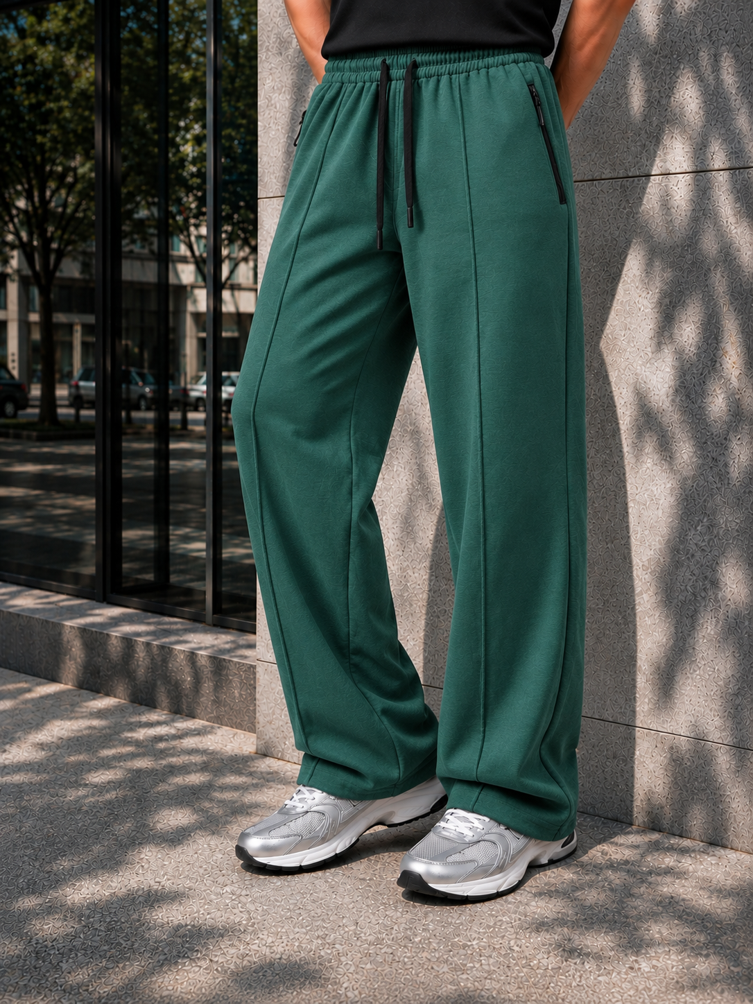 A person wearing dark green wide-leg sweatpants with a black drawstring waist, standing outdoors against a stone wall.