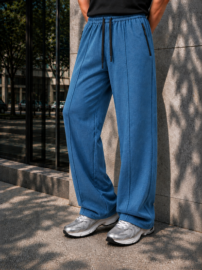 A person wearing wide-leg blue sweatpants with a black drawstring, paired with a black top and silver sneakers, standing outdoors against a stone wall.