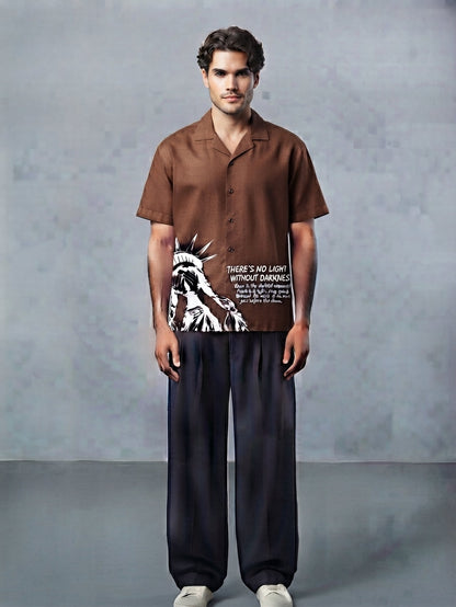 A man stands against a textured gray studio backdrop wearing a brown short-sleeve button-up shirt with a large black-and-white Statue of Liberty graphic and text on the front, paired with dark wide-leg pants and white sneakers.