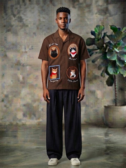 A man stands in a studio wearing a brown short-sleeve button-up shirt with multiple embroidered cat-themed patches, paired with loose dark pants and white sneakers. The background features a textured gray wall and a potted green plant.
