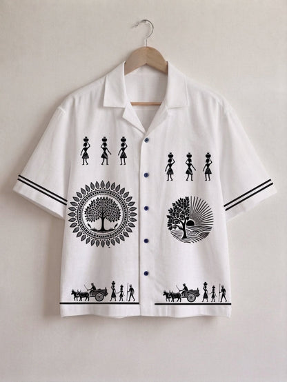 A white short-sleeve button-up shirt with black graphic prints, hanging on a wooden hanger against a light beige wall. The shirt features black silhouettes of figures, circular tree designs, and stripes on the sleeves.