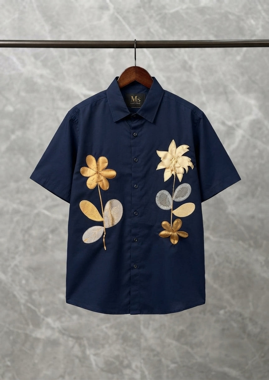 A navy blue short-sleeve button-up shirt hanging on a wooden hanger against a gray marble-patterned wall. The shirt features large, stylized floral embroidery in gold, beige, and light gray on the front.