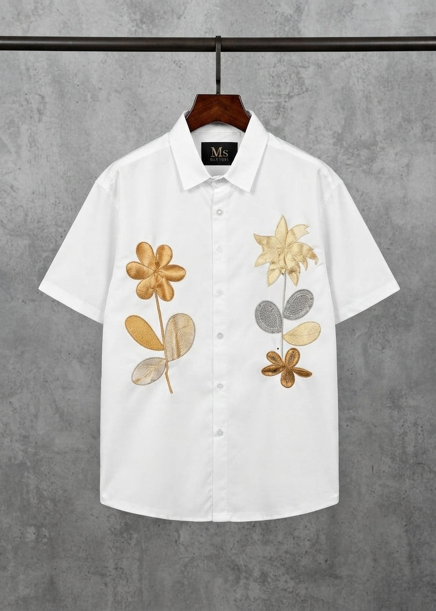 A white short-sleeve button-up shirt with embroidered floral designs in gold, beige, and gray, hanging on a wooden hanger against a textured gray wall.