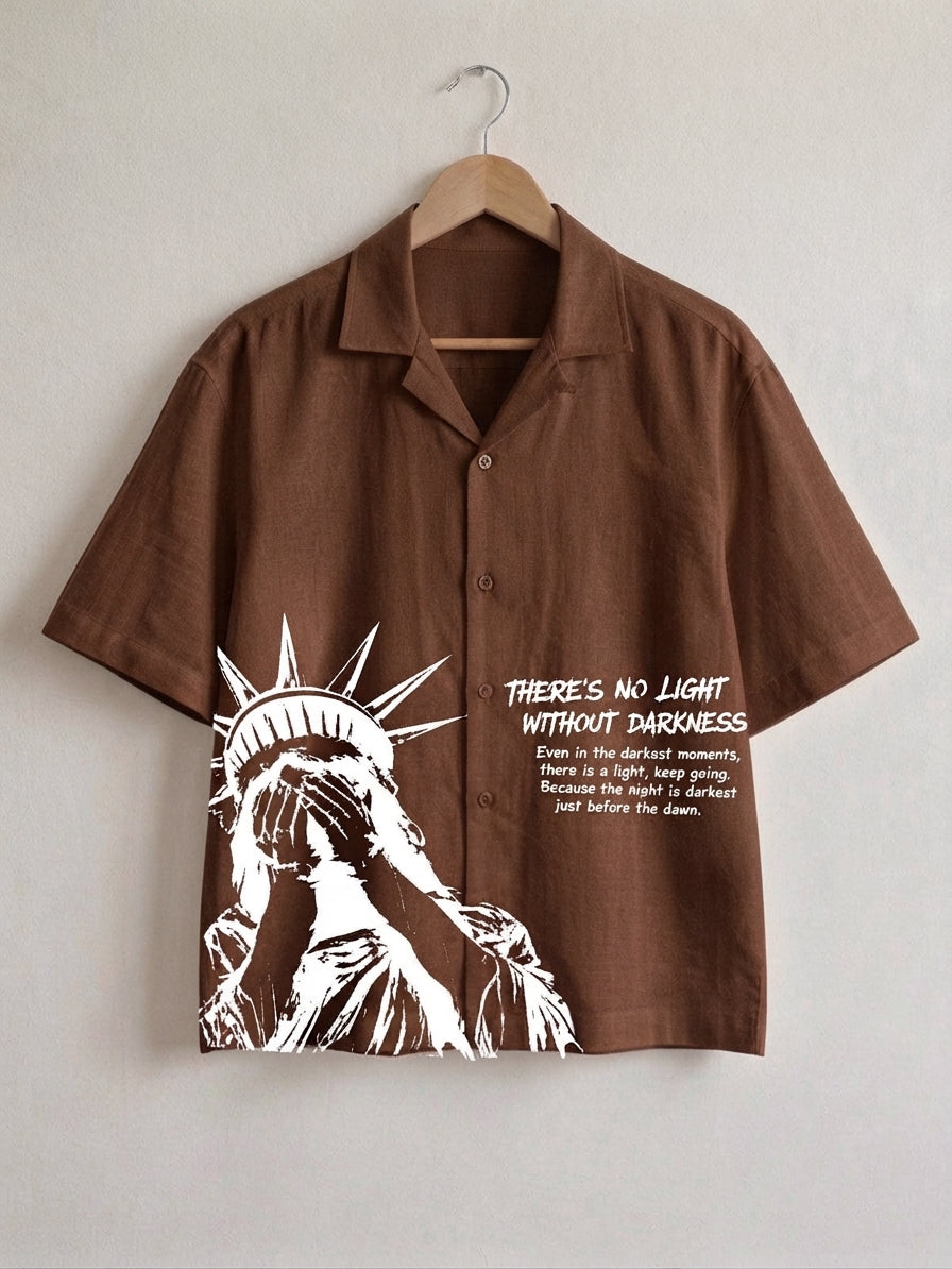 A brown short-sleeve button-up shirt hanging on a wooden hanger against a light gray wall. The shirt features a white graphic of the Statue of Liberty on the left side and white text on the right that reads, 'THERE'S NO LIGHT WITHOUT DARKNESS' with smaller descriptive text below.