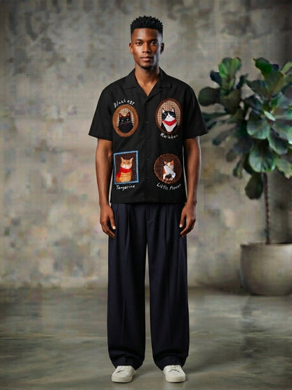 A man wearing a black short-sleeve button-up shirt with four embroidered cat-themed patches on the front, paired with dark wide-leg trousers and white sneakers, standing in front of a textured concrete wall with a potted plant in the background.