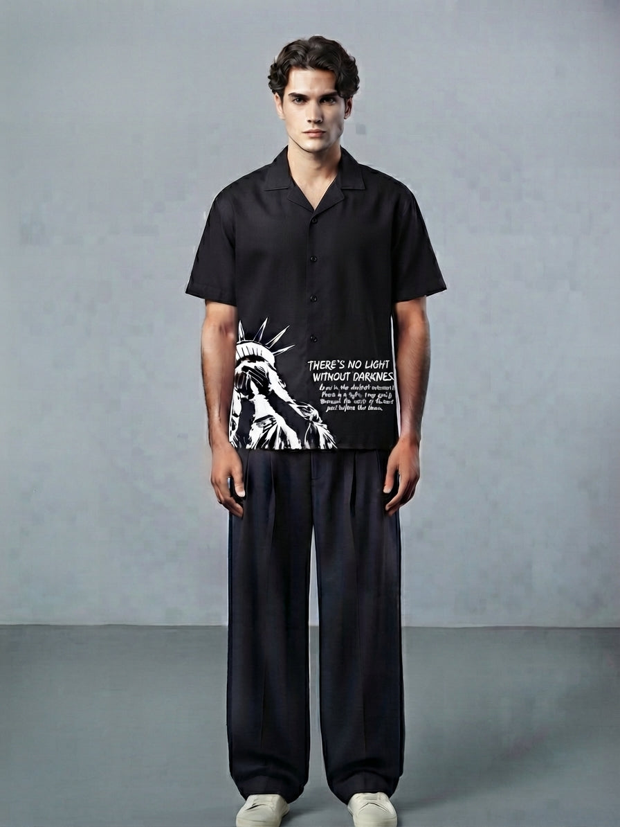 A man stands against a textured gray studio background wearing a black short-sleeve button-up shirt with a white graphic print of the Statue of Liberty and the text 'THERE'S NO LIGHT WITHOUT SURRENDER', paired with loose dark pants and white sneakers.