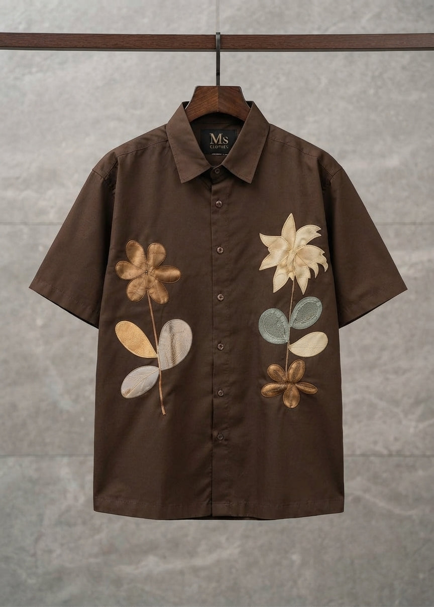 A brown short-sleeve button-up shirt with embroidered tropical floral designs on the front, hanging on a wooden hanger against a textured gray wall.