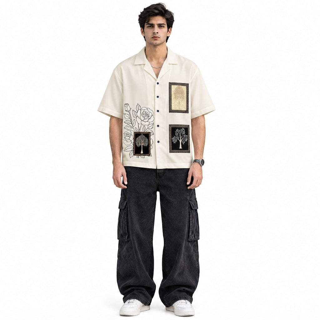 A man modeling a cream-colored short-sleeve button-up shirt with black graphic prints and dark cargo pants, standing against a plain white background.