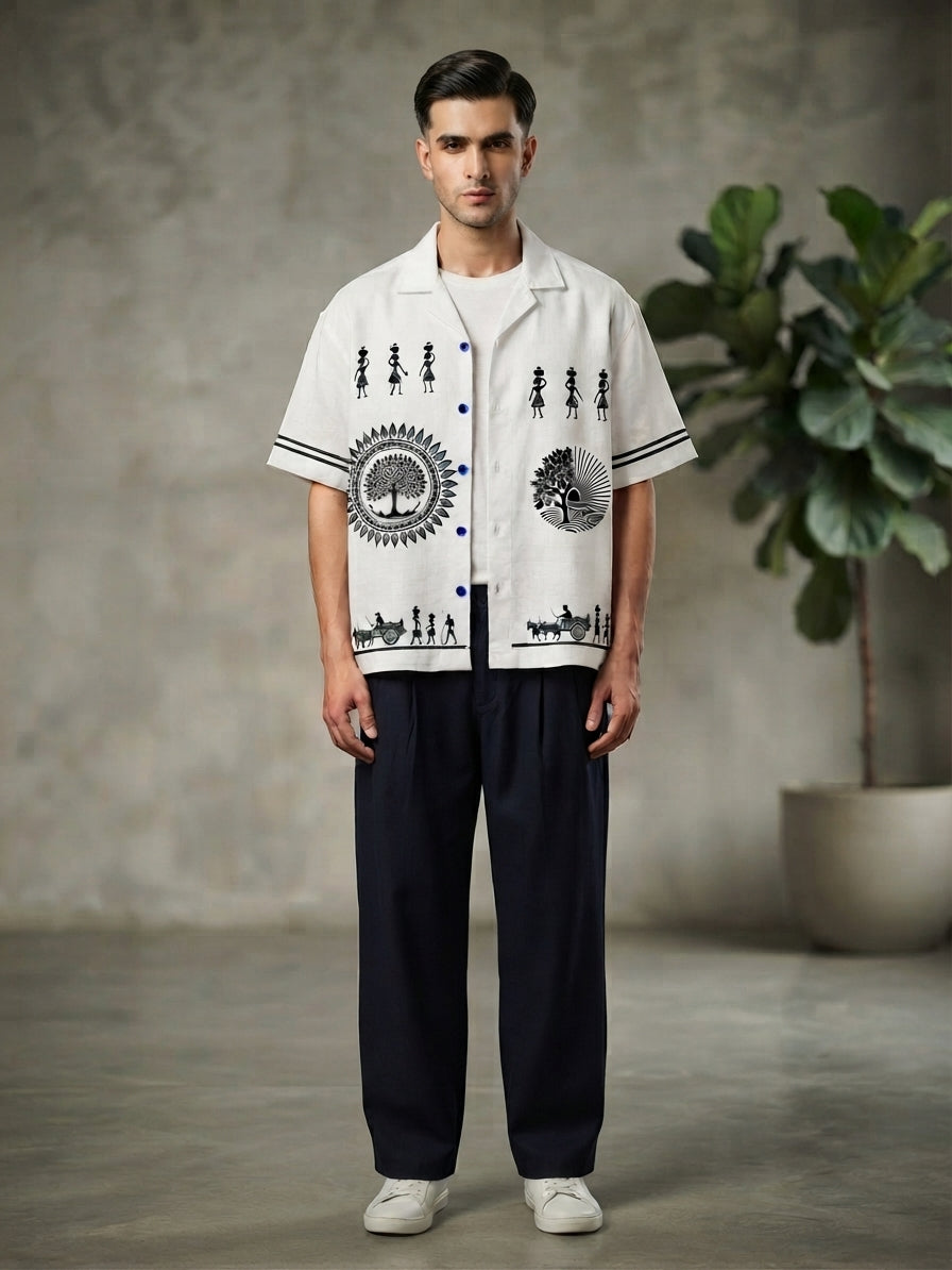 A man stands in a studio wearing a white short-sleeve button-up shirt with black graphic prints, dark navy pants, and white sneakers. The background features a textured gray wall and a potted green plant.