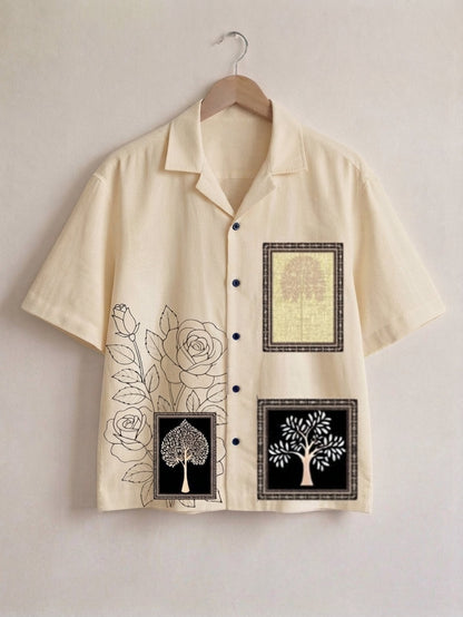 A cream-colored short-sleeve button-up shirt hanging on a wooden hanger against a light beige wall. The shirt features black line art of roses and tree motifs, along with three square graphic panels containing tree illustrations in black, white, and a textured yellow background.