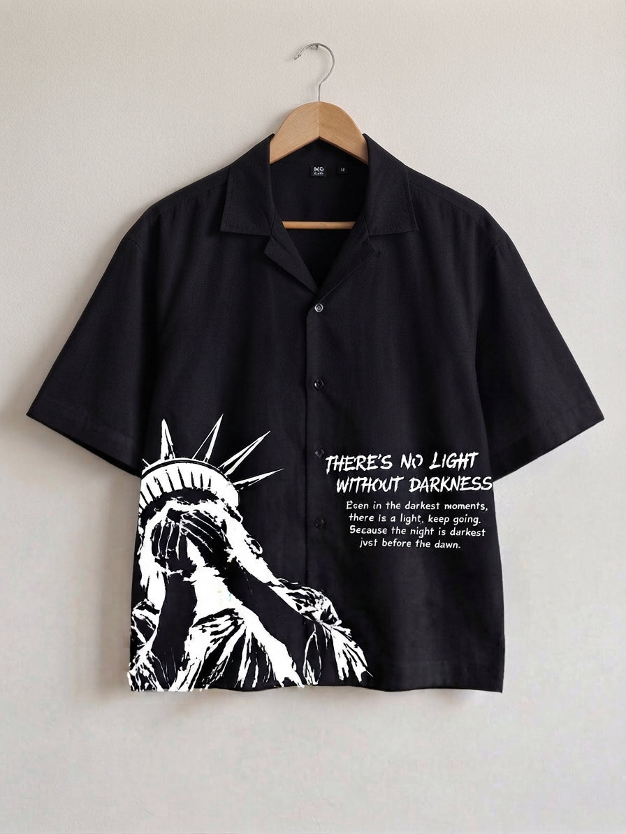 A black short-sleeve button-up shirt hanging on a wooden hanger against a light gray wall. The shirt features a high-contrast white graphic of the Statue of Liberty on the lower left and white text on the right that reads, 'THERE'S NO LIGHT WITHOUT DARKNESS' with a smaller quote below.