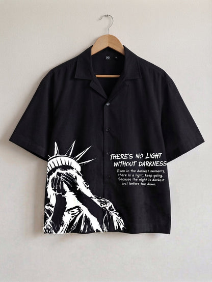 A black short-sleeve button-up shirt hanging on a wooden hanger against a light gray wall. The shirt features a high-contrast white graphic of the Statue of Liberty on the lower left and white text on the right that reads, 'THERE'S NO LIGHT WITHOUT DARKNESS' with a smaller quote below.