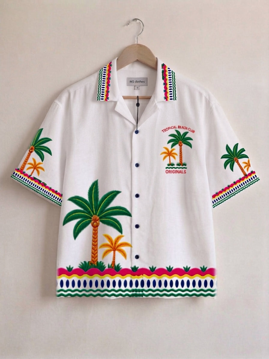 A white short-sleeve button-up camp shirt with colorful tropical palm tree embroidery and patterned trim, hanging on a wooden hanger against a plain beige wall.