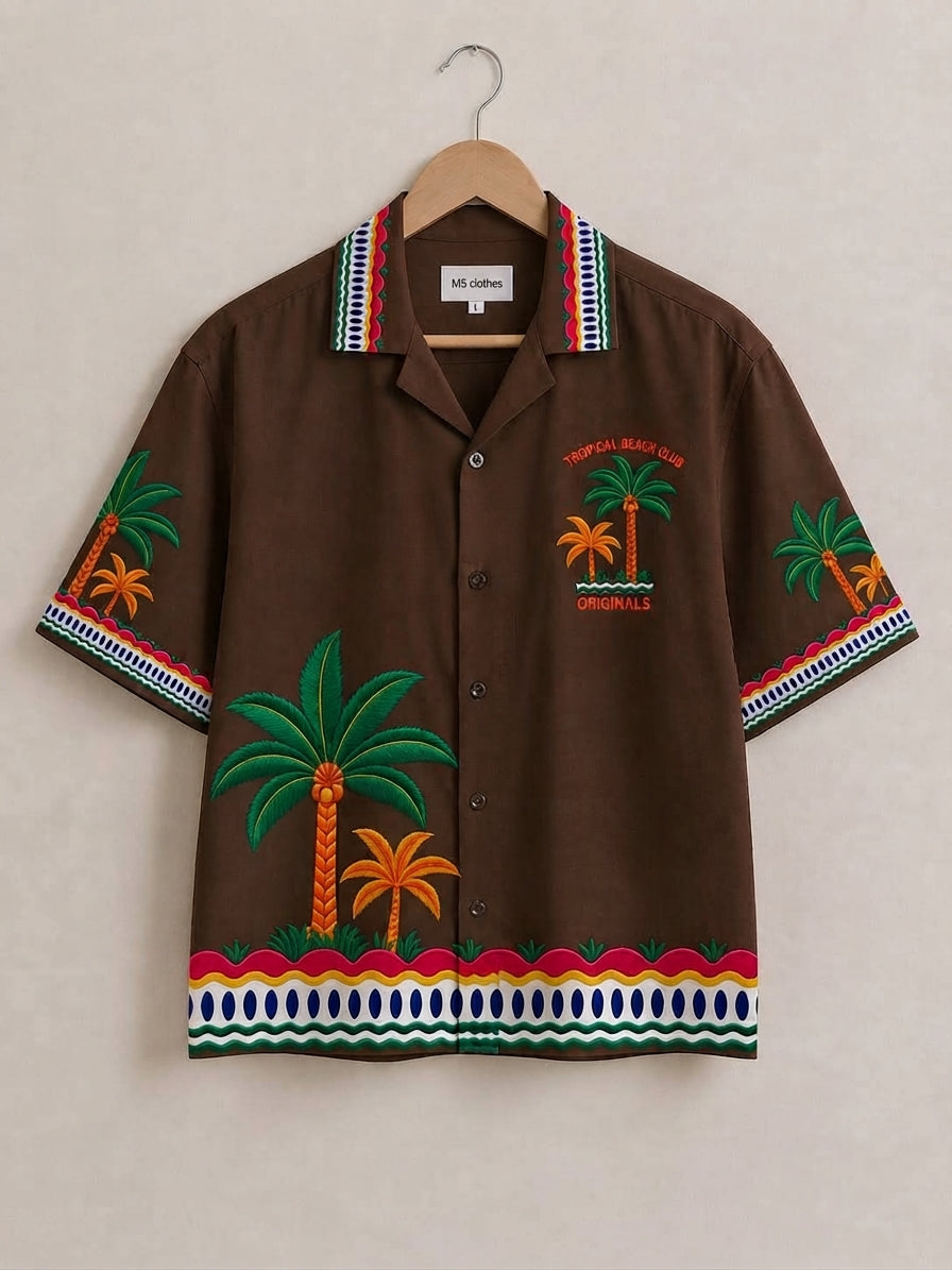 A brown short-sleeve button-up camp shirt with embroidered palm trees and colorful patterned trim, hanging on a wooden hanger against a light beige wall.