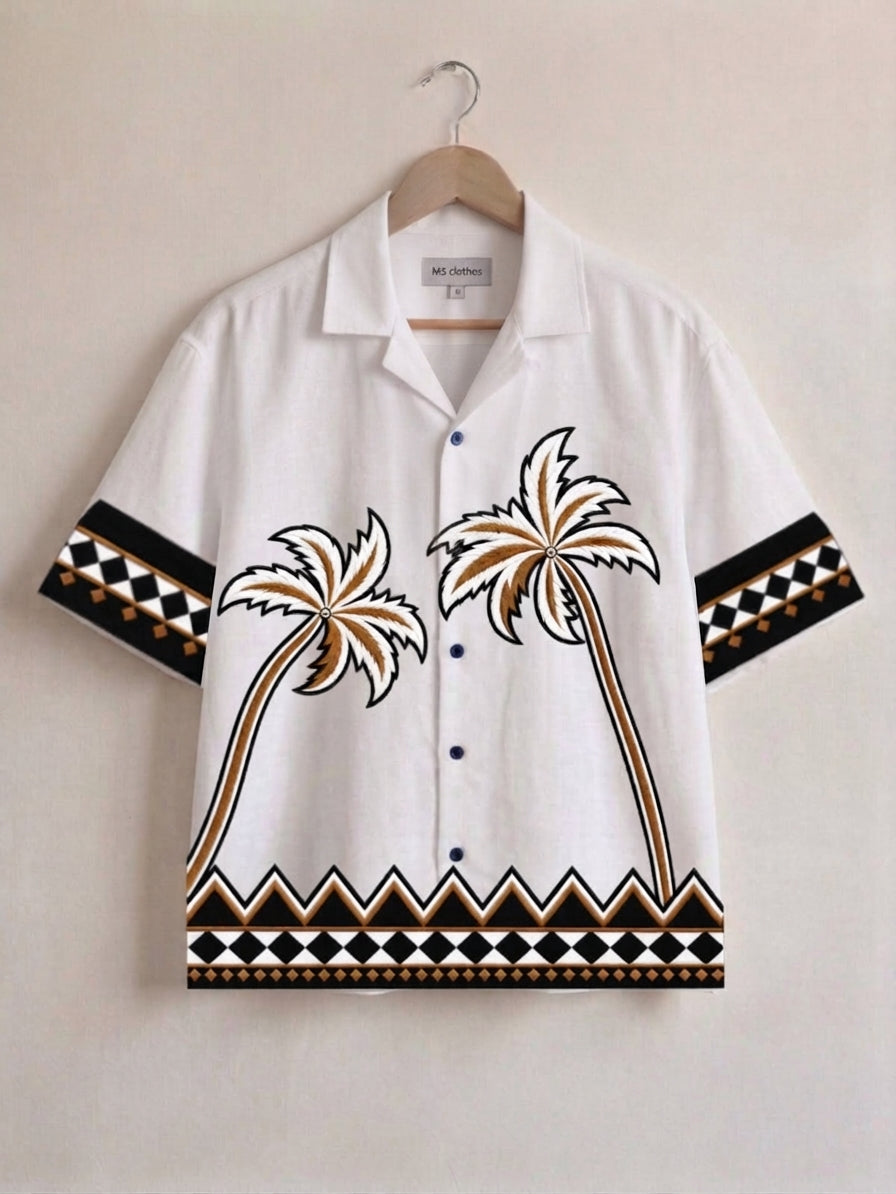A white short-sleeve camp collar button-up shirt with embroidered palm trees and geometric patterns on the sleeves and hem, hanging on a wooden hanger against a light beige wall.