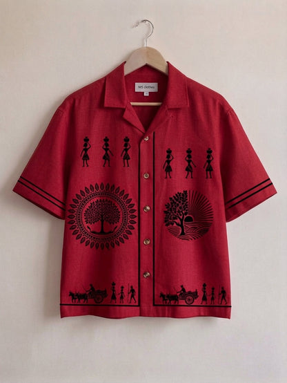 A red short-sleeve button-up shirt with black printed silhouettes of figures, circular tree motifs, and small cart scenes, hanging on a wooden hanger against a plain off-white wall.