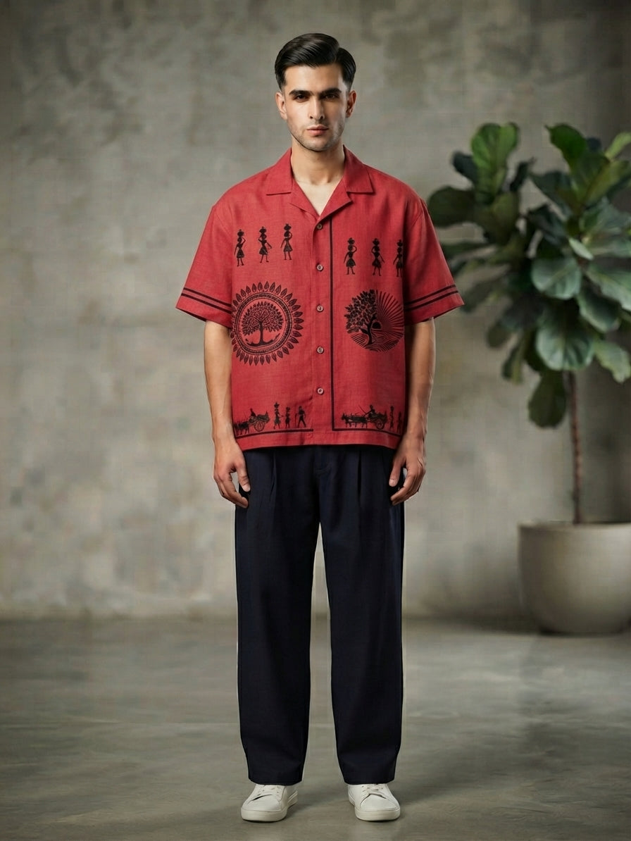 A man stands facing forward wearing a red short-sleeve button-up shirt with black printed graphics and dark navy pants, photographed in a studio with a textured gray wall and a potted plant in the background.