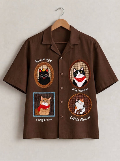 A brown short-sleeve button-up shirt hanging on a wooden hanger against a light beige wall. The shirt features four embroidered cat illustrations with labels reading 'black egg', 'Rainbow', 'Tangerine', and 'Little flower'.