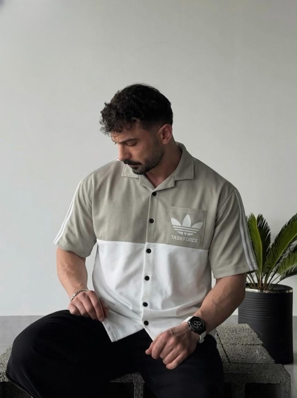 A man wearing a color-blocked short-sleeve button-up shirt by Adidas, featuring a light gray upper section with a chest logo and white lower section, paired with black pants, sitting in a minimalist indoor setting.