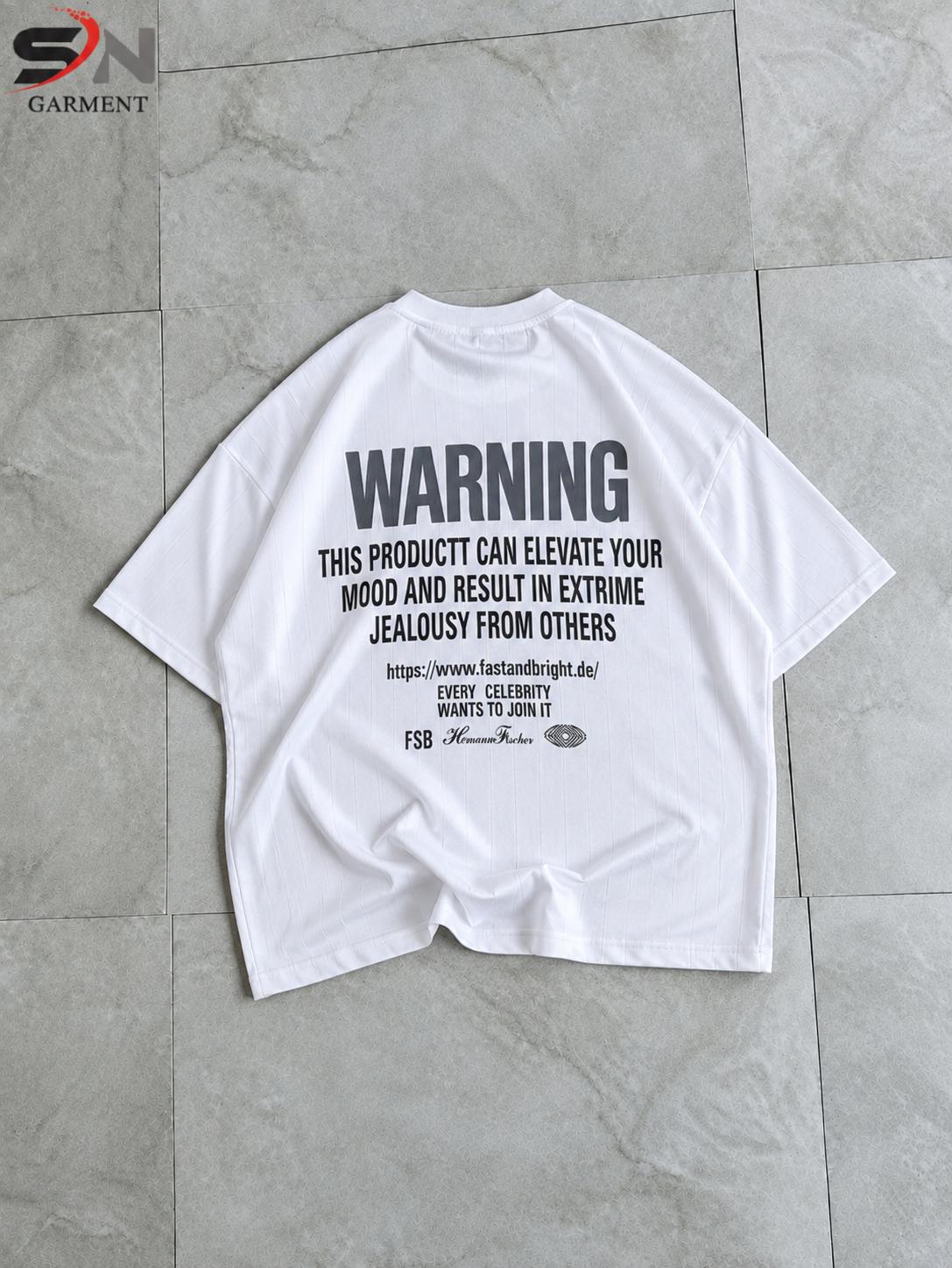 A white oversized graphic t-shirt laid flat on a gray tiled floor. The shirt features a bold black warning text graphic on the front.