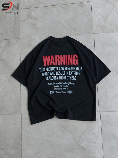A black oversized t-shirt laid flat on a light gray tiled floor, featuring a humorous warning graphic printed on the back in red and white text.