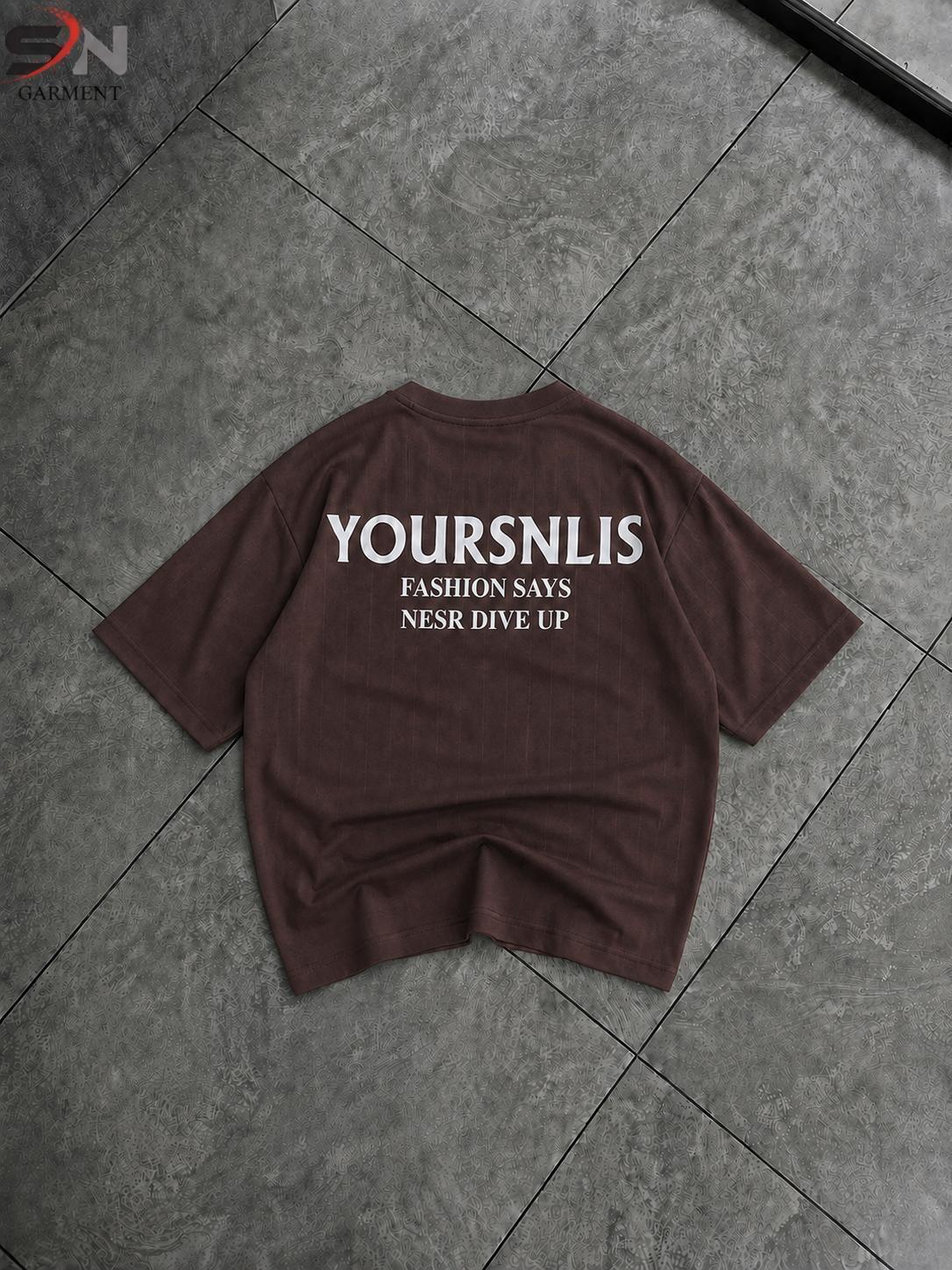 A dark brown short-sleeve t-shirt with white printed text on the back reading 'YOURSNLIST', 'FASHION SAYS', and 'NESR DIVE UP', laid flat on a gray tiled floor.