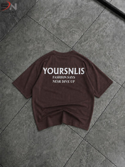 A dark brown short-sleeve t-shirt with white printed text on the back reading 'YOURSNLIST', 'FASHION SAYS', and 'NESR DIVE UP', laid flat on a gray tiled floor.