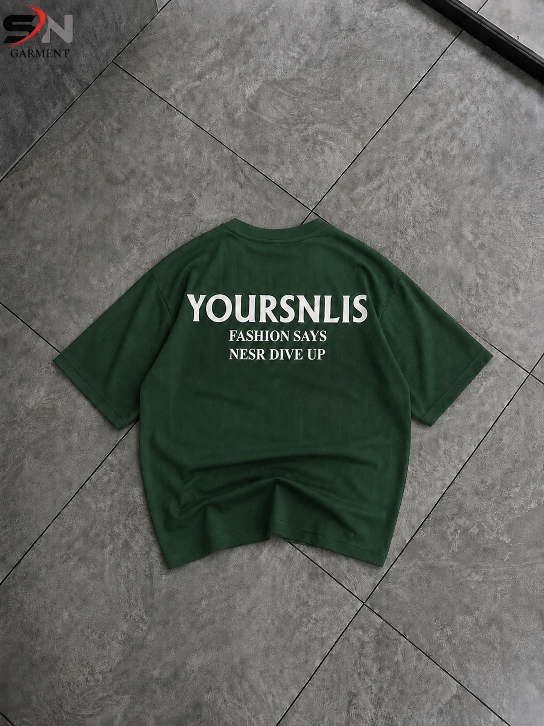 A dark green short-sleeve t-shirt with white printed text reading 'YOURSNLIS FASHION SAYS NSR DIVE UP' laid flat on a gray tiled floor.