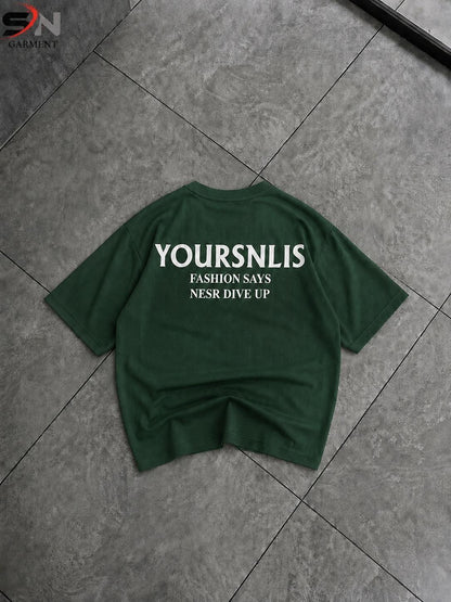A dark green short-sleeve t-shirt with white printed text reading 'YOURSNLIS FASHION SAYS NSR DIVE UP' laid flat on a gray tiled floor.